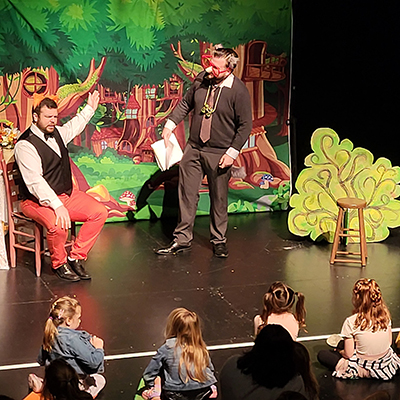 Two costumed men perform in front of a forest backdrop for an audience of children.