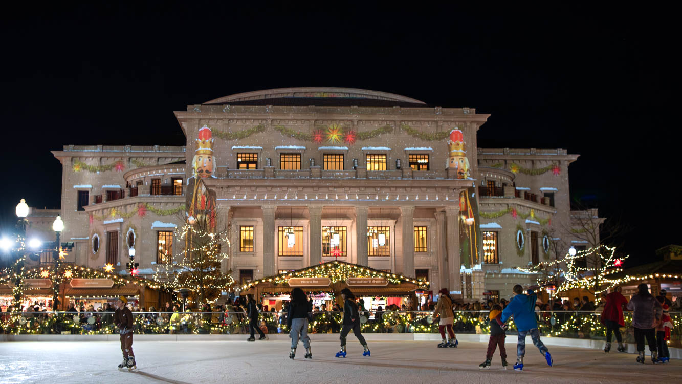 Guests at the Carmel Christkindlmarkt ice skate in front of the Payne & Mencias Palladium with festive lighting.