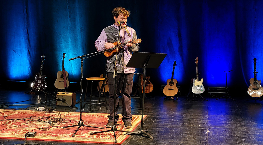 Songwriting 2025 914X504 1 A man performs on ukulele at the Songwriters in the Round performance. Several other guitars sit on stands behind him.