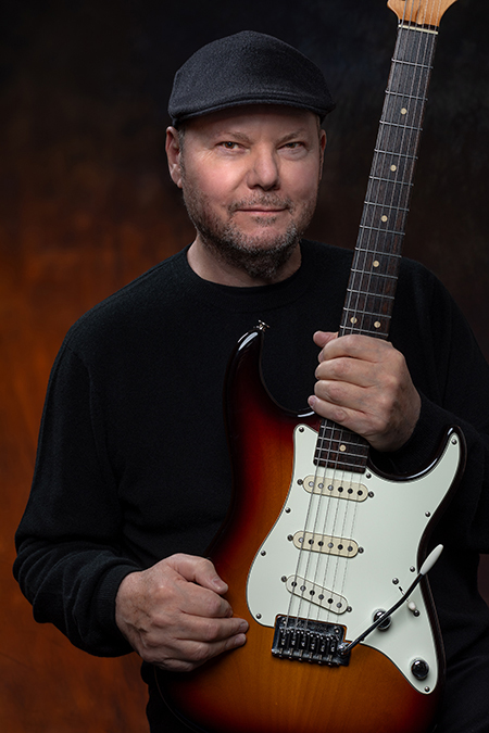 A bearded man in a black sweater and cap holds a Fender Stratocaster electric guitar with a white pickguard and sunburst finish.
