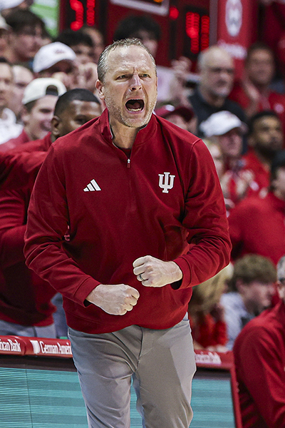 A basketball coach in a red team jacket yells from courtside during a game.