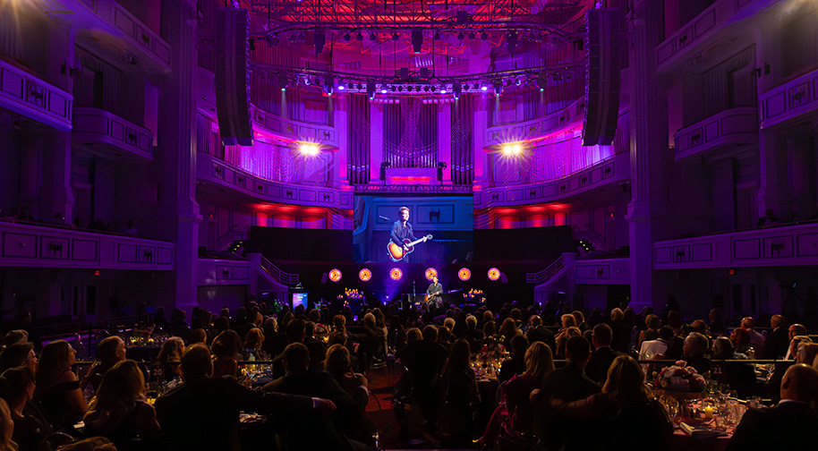Viewed from the back of the concert hall, guests are bathed in warm light as Richard Marx performs on guitar against blue lighting and candles.