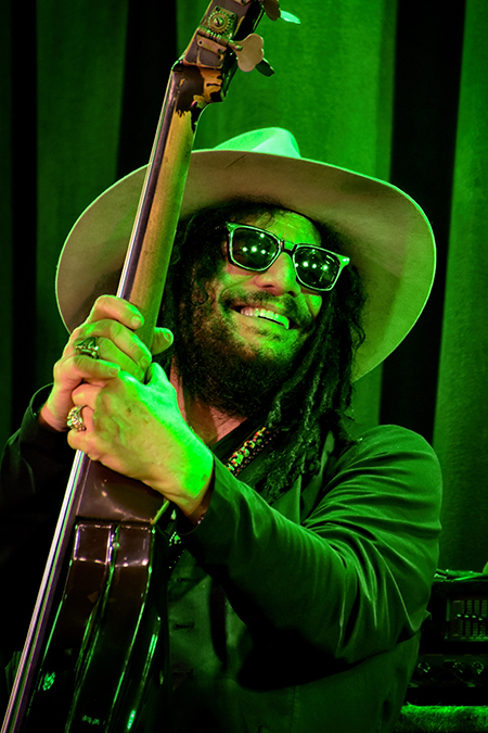 A bearded man with dark glasses and a wide-brimmed hat smiles while gripping an upright bass.