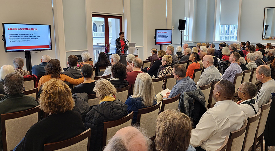 A woman presents a talk at a Luminaries event with screens displaying information about spiritual music.