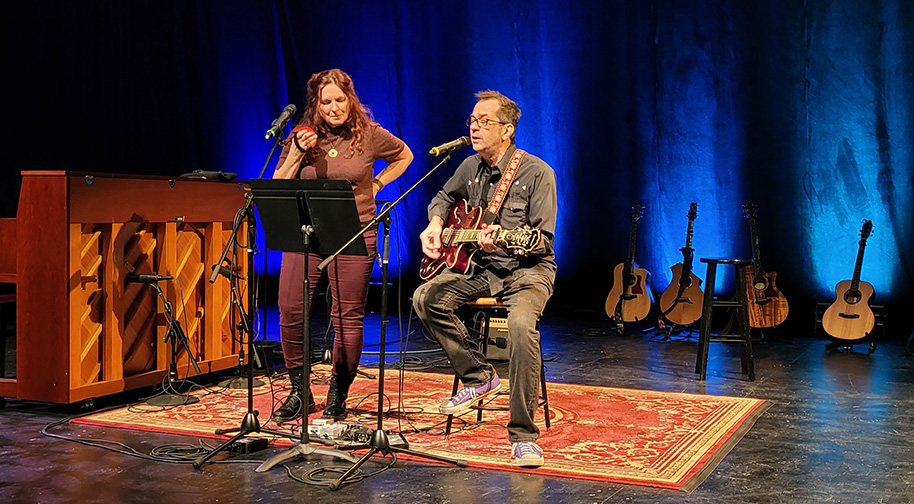 Songwriting 2025 914X504 3 A man sits and plays guitar while a woman plays a shaker and sings.
