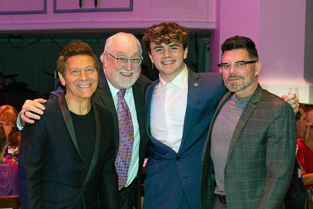 Three men and a teen boy wearing suits smile as they pose for a photo on a stage.
