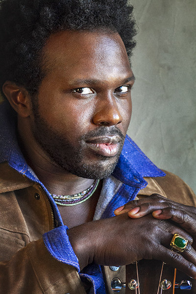 A black man in a blue shirt and suede jacket looks at the camera with hands clasped and a mysterious smile.
