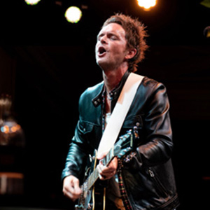 A man wearing a leather jacket plays an electric guitar on a stage.