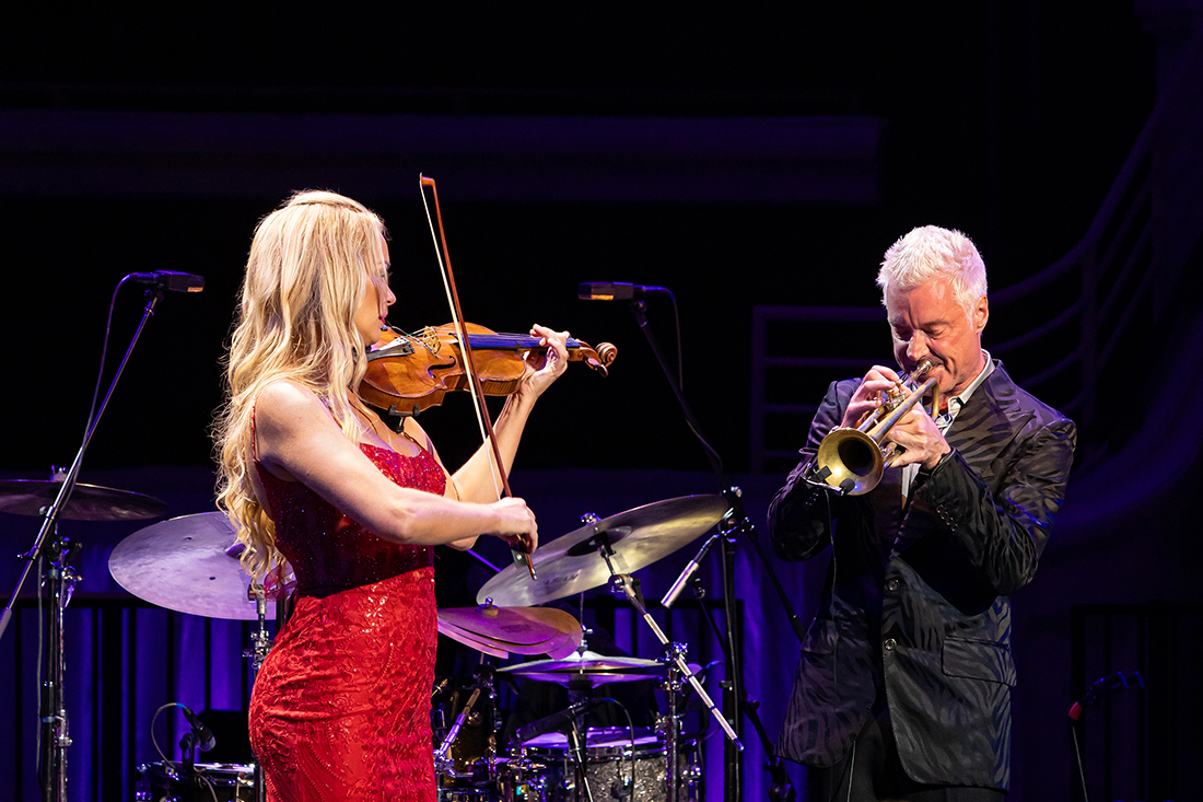 A female violnist in a red dress and a male trumpeter in a suit perform onstage.