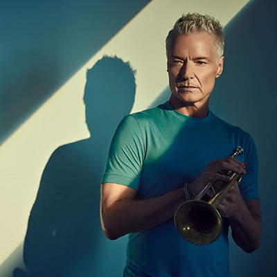 A man in a blue T-shirt, musician Chris Botti, holds a trumpet as he looks into the distance.