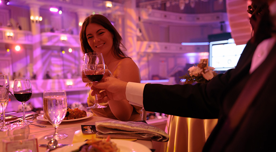Guests at a gala event toast with their wine glasses at a table in a box.