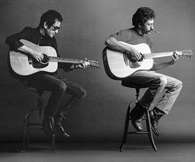A black and white photo shows two men sitting on stools playing acoustic guitars.