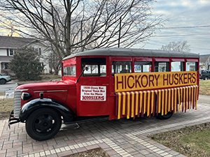 An old red school bus has a yellow banner on the side reading "Hickory Huskers."