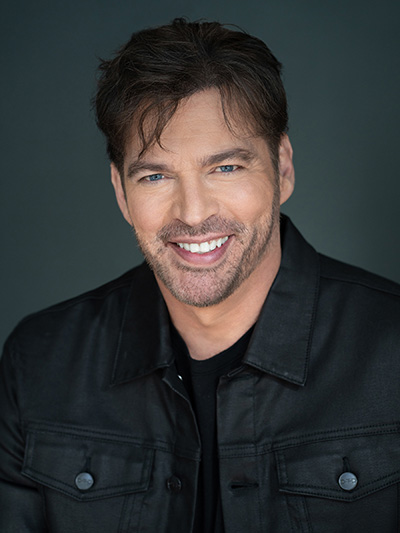 A man with tousled dark hair and stubble smiles while wearing a black jacket and T-shirt.