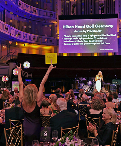 In a crowded, glittery ballroom, a woman raises a yellow paddle to bid in an auction as another woman on stage reacts.