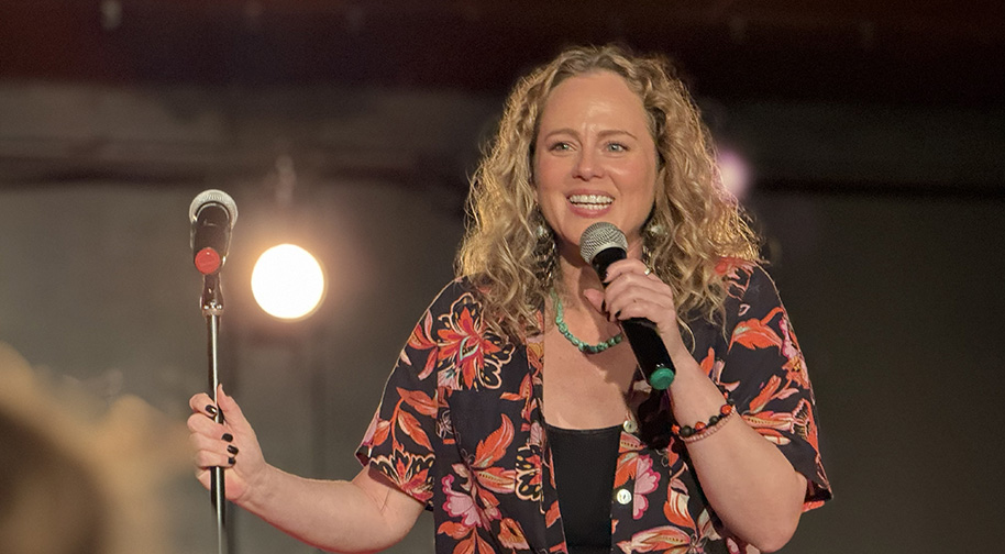 Lasley 914X504 Comedian Sherra Lasley performs at a comedy club under a spotlight.