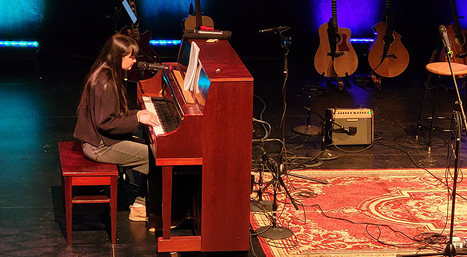 Songwriting 2025 914X504 4 A woman plays piano and sings at the Songwriters in the Round performance.