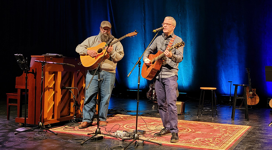Songwriting 2025 914X504 5 Two men stand to play acoustic guitar and sing side by side.